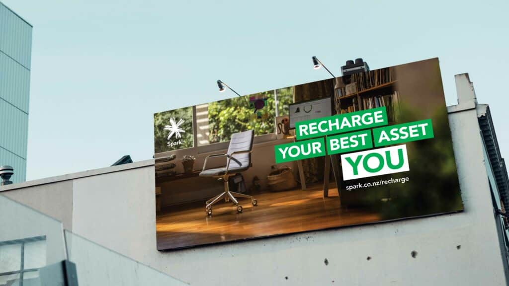 A photograph of a Spark billboard on the side of a building. The billboard says "Recharge your best asset. You. spark.co.nz/recharge"