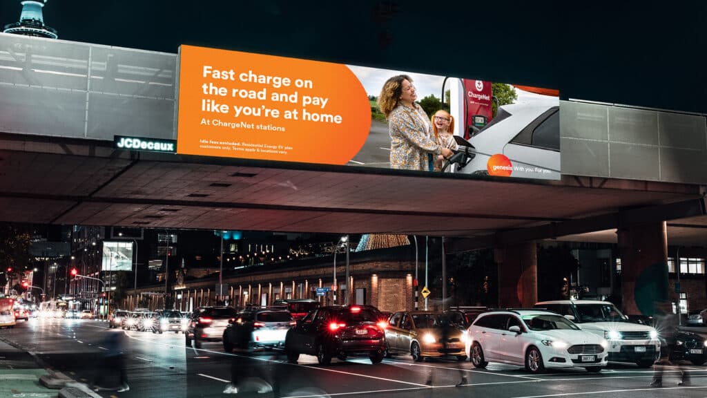 A photograph of a Genesis billboard above a highway. The billboard says "Fast charge on the road and pay like you're at home"