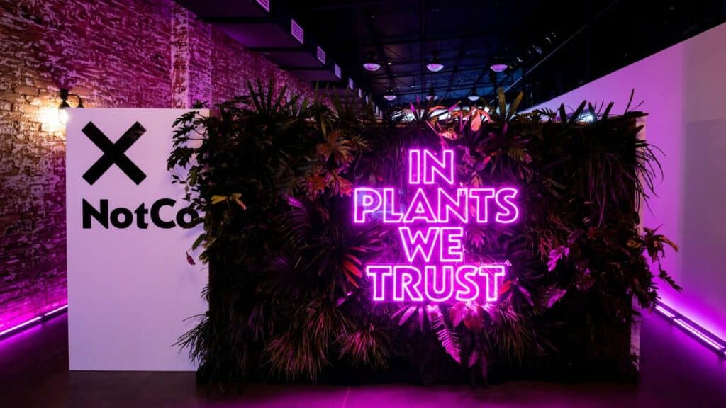 A photograph of a wall decorated with plants and the words "In plants we trust" are displayed on top of the plants with the NotCo logo displayed next to it